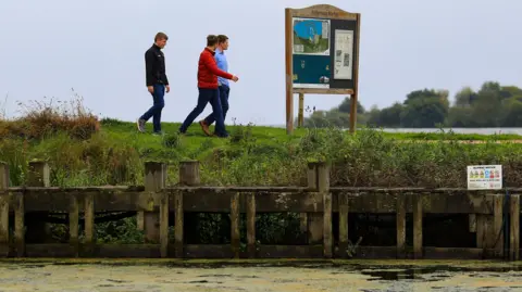 Three men walking next to Lough Neagh, which has a layer of blue-green algae on the surface