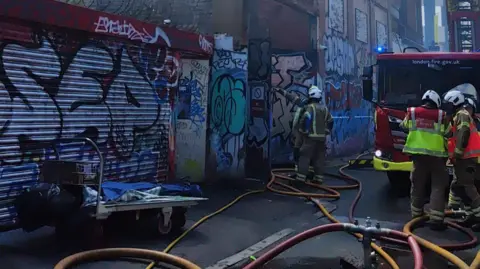 London Fire Brigade A fire truck and firefighters stand by hoses in a back street in London. There is graffiti on the walls on one side of the road.