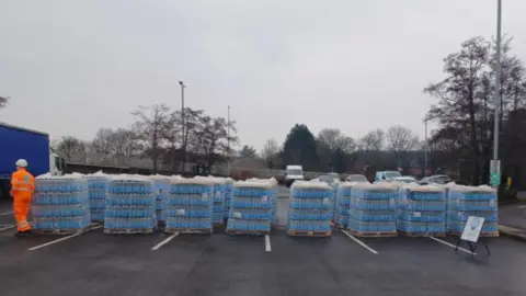 Thames Water Around 20 wooden pallet crates stacked high with bottled of water, wrapped and sealed in clear plastic. They are sitting in the middle of a car park on a grey and dreary day. On the left there is a Thames Water employee dressed in full hi-vis orange with a white plastic helmet.