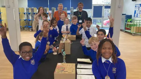 Pupils from Camp Hill Primary School in Nuneaton are wearing blue jumpers and white shirts are smiling at the camera and are making the 'V for Victory' sign, while sitting alongside the art they have created.