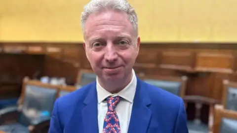 Alex Allinson standing in the Tynwald chamber looking straight ahead smiling. He is wearing a blue suit with a white tie and a blue, white and red tie.