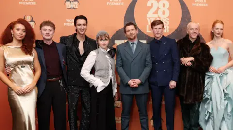 Sony Pictures UK Eight of the 28 Years Later cast members stand in front of a photo wall at the premier. Locke wears a burgundy shirt, navy blazer and black trousers with a brown belt. O'Connell wears a grey shirt, with a darker grey suit jacket, trousers and tie. 