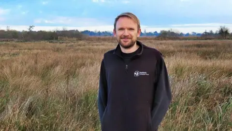 Suffolk Wildlife Trust Lewis Yates standing in a grassy field. He is wearing a black fleece and is smiling at the camera. He has brown hair and a beard.