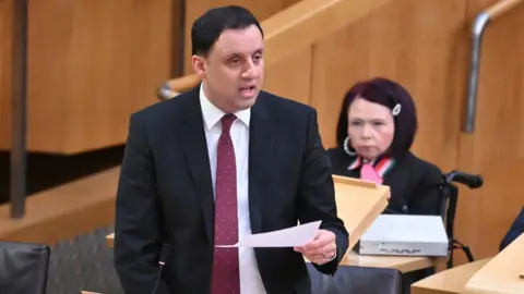 Getty Images Anas Sarwar, who has short black hair, speaks at a podium with a piece of paper in his left hand. He is wearing a dark suit, white shirt and red tie. 