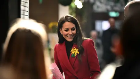 Ben Stansall/PA Wire The Princess of Wales is smiling as she looks down, she is wearing a red coat and is holding a pink and white polka dot tube. Catherine has a large yellow daffodil broach attached to her red coat. She is in focus and the foreground is people waiting to meet her, their heads are out of focus.
