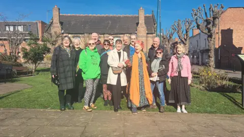 13 people huddled together for their photograph to be taken. The people are wearing a mixture of clothing. They are standing on the edge of a small lawn in a town centre, with a large bricked building with two chimneys behind them.