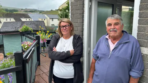 Julie and Daryl Wilkins stand outside Daryl's house, Julie with her arm crossed. He is leaning against the wall with the front door open beside him. Julie has chin-length blonde hair and is wearing glasses, a white t-shirt and a black jacket. Behind them decking can be seen with flower planters, with a row of terraced houses in the background.