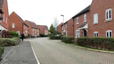 A street of houses on Great Western Park, which is a large estate in Didcot. The homes are red bricked with hedges out the front. A man walks away from the camera on the pavement on the left. 