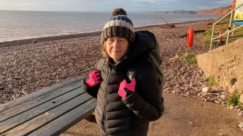Sea swimmer Sarah Giles is standing on Budleigh Salterton beach. She is wearing a black coat, pink gloves and a brown, white and blue stripey woolly hat with a bobble. She is standing next to a wooden bench. The beach is pebbly. There are cliffs in the distance.