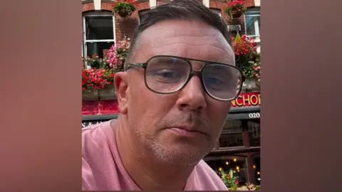 Head and shoulders shot of Graeme Hull. He is wearing glasses and had short dark hair. He is wearing a pink t-shirt.