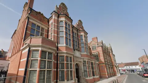 The photo shows a red-brick Victorian-style building with large bay windows, stone detailing and an arched entrance. It stands on a corner in a town with nearby shops and cars.