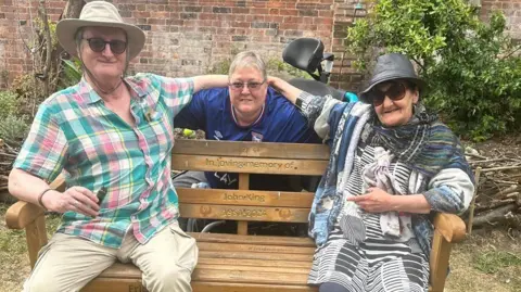 Cad Taylor, BBC Two people sit on a bench and someone is in the middle from behind the bench, there is an inscription on the bench, in loving memory of John King, 1951-2024