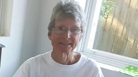 Supplied Suzanne Rees, wearing a white t-shirt and black wire glasses smiles in a photo