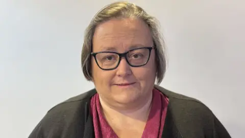 The picture shows a person wearing glasses, a pink top and a dark cardigan, standing indoors against a plain light-coloured wall.