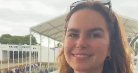 Molly smiling at the camera. She has her sunglasses resting at the top of her head and in the background there's a white canopy and some crowds lining what looks like a racetrack.