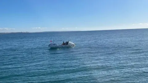 Rock PR Zara Lachlan en un barco blanco de manga ancha de 7 m de largo. Ella rema en las tranquilas aguas azules del océano, con un cielo azul más pálido arriba.