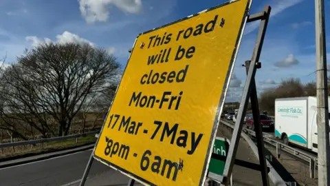 Andrew Turner/BBC A yellow road sign, stating: This road will be closed Monday to Friday 17 March to 7 May, 8pm to 6am. In the backdrop are trees, a white clouded blue sky, and a queue of traffic including a van bearing the name "Lowestoft Fish Co."