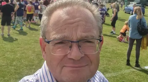 Jeremy Pert Jeremy Pert wears glasses and a striped shirt, he's in a field surrounded by people. It's a sunny day. He's out campaigning.