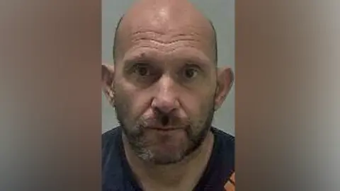 West Mercia Police Blurry mugshot of a bald man with brown eyes wearing a navy top