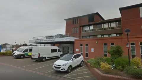The outside of the reception area of The Friarage Hospital. The building is made of brick and there are an ambulance, a white van and a white car parked outside.