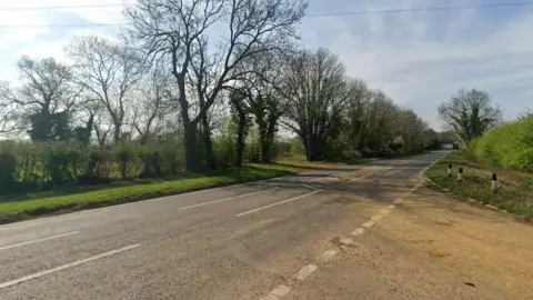 Google A Google Street View looking at Newington Road, an A-Road, from the Bloxham Road. Tall trees run along one side of the road, a field is beyond.