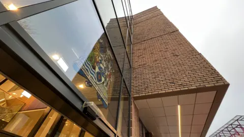 A picture of a modern building with a glass front and a coat of amrs on it.