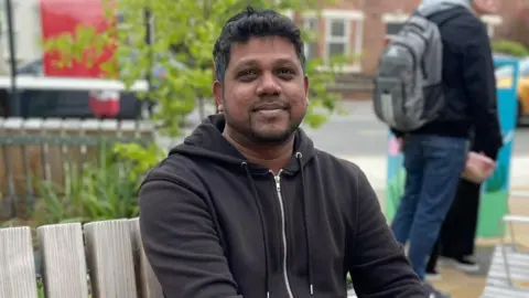 Tom MacDougall/BBC Thameshwar Kheran, a man in his mid-30s of South Asian heritage, sitting on one of the benches in the garden.