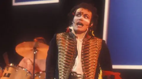 Adam Ant on stage in the 1980s with a drum kit behind him. He is wearing a gold military-style jacket with lots of buttons and brocade and has his traditional white stripe painted across his nose and upper cheeks. He appears to be mid song.