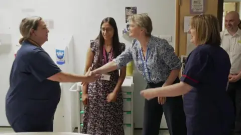 Several hospital staff members gathered in a ward or clinical area. One staff member is shaking hands with Yvette Cooper while colleagues stand nearby. Medical storage units, a doorway and clinical equipment are visible in the background.