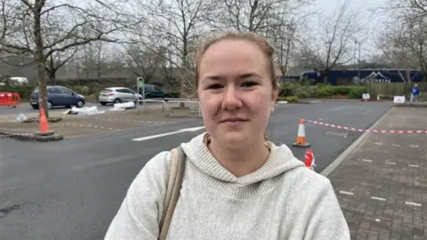 A woman with her hair back in a ponytail wears a cream hoodie and looks at the camera. She is standing in a car park that has been taped off and turned into a bottled water station.