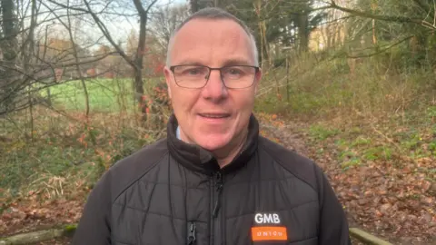 A man is wearing a black coat that says GMB Union on it. He wears glasses. Trees are behind him. 