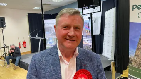 Dennis Jones smiling and looking at the camera. He is wearing a blue blazer, white shirt and red Labour rosette. In the background is the stage where local election results are announced. 