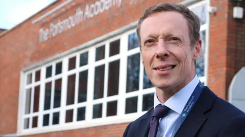 Sam Poole Headteacher Dan Botting standing outside The Portsmouth Academy. He is wearing a suit, blue shirt and purple tie, and smiling