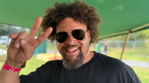 A man with dark hair and beard and wearing sunglasses and a black t-shirt smiling underneath a green gazebo with his right hand making the peace sign using two fingers 