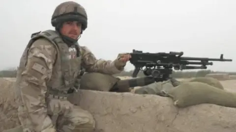 Family photo Matthew Evans in a soldier's fatigues and a machine gun on sand bags.