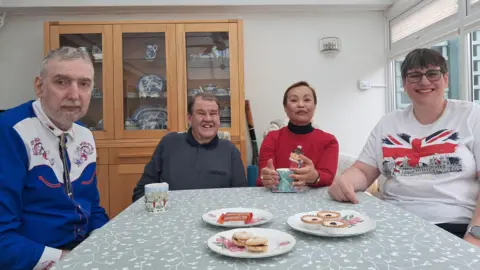 Four people sit around a dining table laden with biscuits and cups of tea.