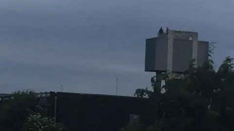 JACOB MAVIN A blurry photograph of a rectangular, concrete water tower on grey stilts with two human figures standing on the top. There is tree foliage and metal fencing in the foreground.  