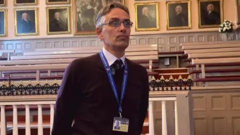 Rob Ward, Barnstaple Town Council Clerk stood in a room with lots of wooden benches behind him. He is looking away from the camera with a stern look on his face. 
