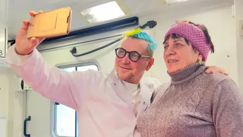 A doctor smiles as he takes a selfie with a woman patient inside a mobile medical unit. His hair is dyed the blue and yellow of the Ukrainian flag, and medical paraphernalia is in the background.