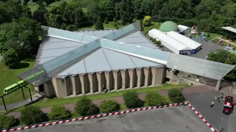 BBC Drone view of a square buiding with a cross design on the toof, with a green monorail emerging from one side and a vintage red bus parked outside on the other side