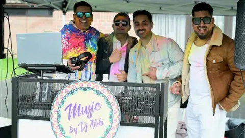 Amith Nair Four men smiling for a picture by a DJ booth with a sign Music by DJ Abhi. The Dj is wearing a multicolour top and is holding a pair of headphones.