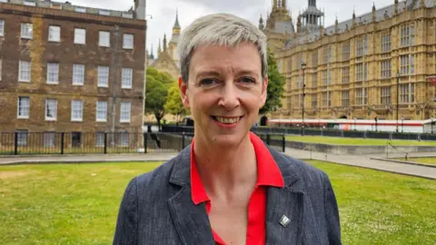 Polly Billington MP is wearing a red shirt and grey jacket. She is standing on Westminster Green with the Houses of Parliament in the background