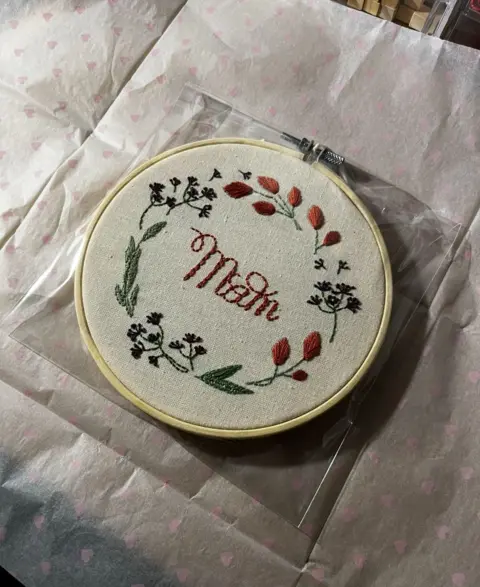 WithYou A piece of embroidery with flowers round the outside and the word "Mam" in the middle