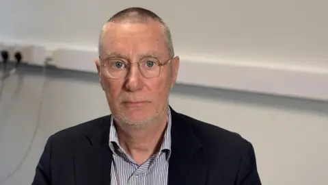Anthony May pictured in his office at the hospital trust. He is wearing a black jacket striped shirt