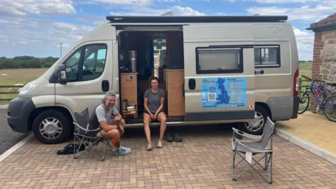 Katie Radley/BBC A woman is sitting in the open door of a campervan, smiling at the camera. On the side of the campervan is a blue poster with a map of Great Britain on it. In front of the campervan, a man is sitting in a camping chair smiling at the camera. There is an empty camping chair on the right hand side. The campervan is parked in a car park. There is a field with sheep behind it.
