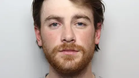 A man with brown/auburn hair and a short ginger beard stands against a grey background