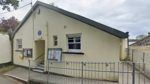 Google Maps Instow Village Hall. It is a single-storey building painted pale yellow that has two windows. There is a ramp running past the front door and leading to the building. There are steps leading to the front door. ON the outside of the building are two noticeboards and a defibrillator.