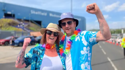 BBC/Nicola Rees Rob and Nikki Brookes. They are dressed in bright blue shirts with Hawaiian-style flowers around their necks. They have their fists in the air in celebration. 