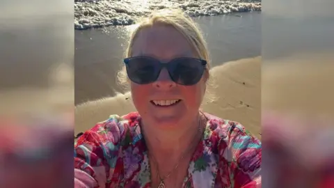 A woman takes a selfie on a beach. It is sunny. The waves are small and gently lap on the beach. 