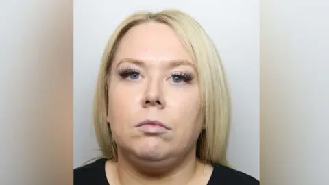 A mugshot of Chelsea Kemp. She has blonde shoulder length straight hair and blue eyes, with false eyelashes. She wears a black top and is staring at the camera. 